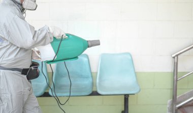 COVID-19 'u önlemek için ofisin dezenfekte edilmesi, ofiste dezenfekte edilmiş beyaz tehlikeli madde giysisi giyen kişi koruyucu maske takıyor ve Coronavirus püskürtüyor..