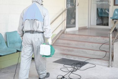 COVID-19 'u önlemek için ofisin dezenfekte edilmesi, ofiste dezenfekte edilmiş beyaz tehlikeli madde giysisi giyen kişi koruyucu maske takıyor ve Coronavirus püskürtüyor..