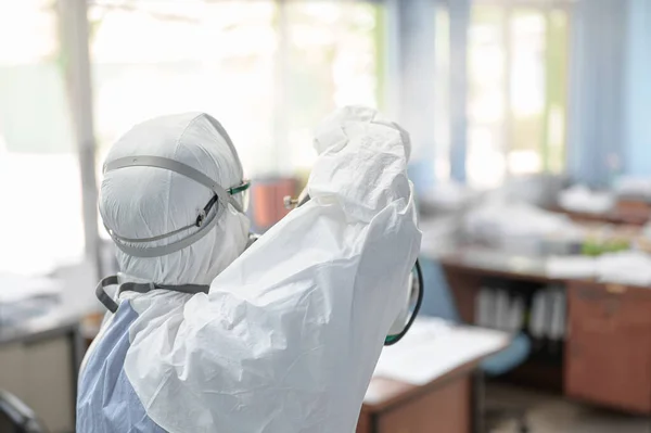 COVID-19 'u önlemek için ofisin dezenfekte edilmesi, ofiste dezenfekte edilmiş beyaz tehlikeli madde giysisi giyen kişi koruyucu maske takıyor ve Coronavirus püskürtüyor..
