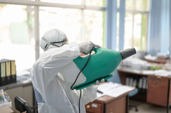 COVID-19 'u önlemek için ofisin dezenfekte edilmesi, ofiste dezenfekte edilmiş beyaz tehlikeli madde giysisi giyen kişi koruyucu maske takıyor ve Coronavirus püskürtüyor..