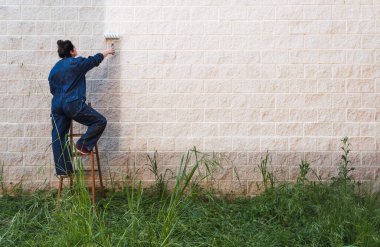 Duvarı eski ahşap bir merdivenin üstüne boya silindiriyle boyayan kadın bir ressam. Etiketlenecek beyaz bir işaret bırak.
