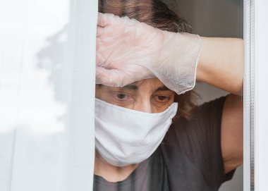 Koronavirüs, covid-19 ya da başka bir hastalığa karşı maske takan üzgün, endişeli kadın karantinadayken pencereden bakıyor ve dışarı çıkamıyor.