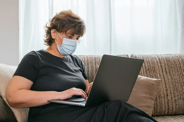 Yalnız bir kadın, Coronavirus 'a karşı bir maske ile korunuyor, covid-19 ya da başka bir hastalığa karşı, evde bilgisayarla çalışıyor, kanepede oturuyor. Kadın oturma odasında dizüstü bilgisayarla çalışıyor. Koronavirüsten korunuyor.
