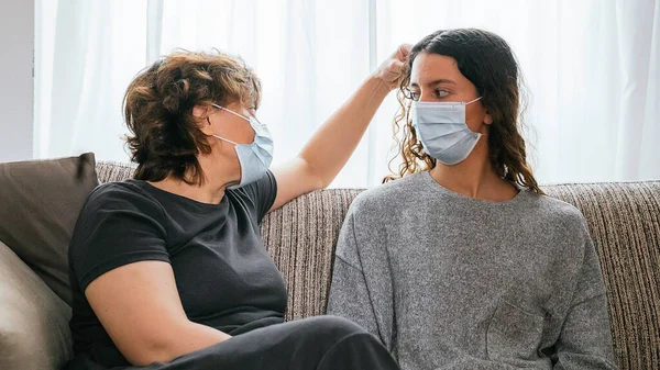 Mother and daughter protected by a mask against coronavirus, covid-19 or any other disease, sitting in their living room talking to each other while the mother caresses her daughter