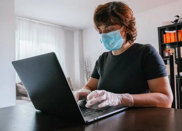 Yalnız bir kadın, eldiven ve maskeyle Coronavirus 'a karşı korunuyor, covid-19 ya da başka bir hastalığa karşı, evden bir dizüstü bilgisayarla çalışıyor, masada oturuyor.
