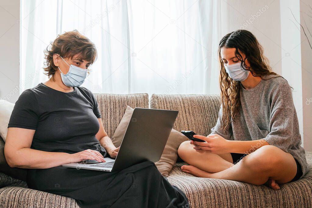 Mother and daughter protected with a mask against coronavirus, covid-19 or any other disease, sitting in their living room using a phone and a computer for work