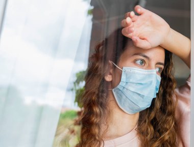 Coronavirus. Koronavirüslü genç bir kadın pencereden dışarı bakıyor ve kendini korumak ve evdeki hastalıktan kurtulmak için maske takıyor. Salgın