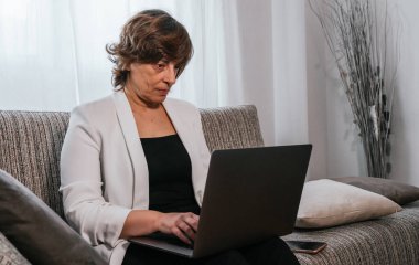 Evdeki bilgisayarını kullanan bir iş kadını. Evden çalışan bir iş kadını. Kanepedeki kadın. Yaşlı kadın çalışıyor. Yaşlı kadın laptopunu kullanıyor. Evden çalışıyorum. İş kadını kanepede oturuyor.