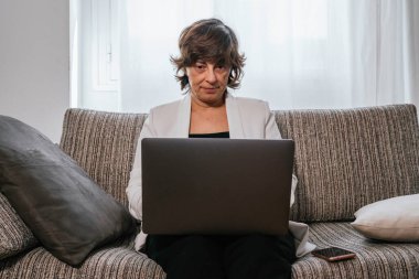 Evdeki bilgisayarını kullanan bir iş kadını. Evden çalışan bir iş kadını. Kanepedeki kadın. Yaşlı kadın çalışıyor. Yaşlı kadın laptopunu kullanıyor. Evden çalışıyorum. İş kadını kanepede oturuyor.