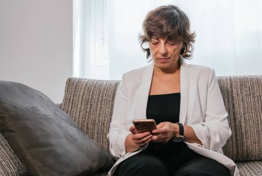 Evde akıllı telefonunu kullanan bir iş kadını. Evden çalışan bir iş kadını. Kanepedeki kadın. Yaşlı kadın çalışıyor. Akıllı telefonu kullanan kıdemli bir kadın. İş kadını kanepede oturuyor.