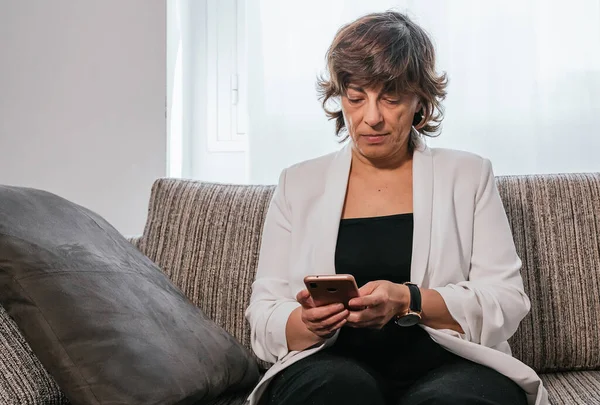 Evde akıllı telefonunu kullanan bir iş kadını. Evden çalışan bir iş kadını. Kanepedeki kadın. Yaşlı kadın çalışıyor. Akıllı telefonu kullanan kıdemli bir kadın. İş kadını kanepede oturuyor.