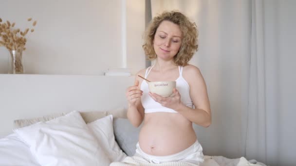 Щаслива вагітна жінка їсть зернові для сніданку в її спальні . — стокове відео