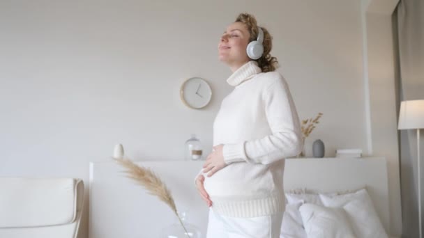 若いです妊娠中の女性身に着けていますヘッドフォンと居心地の良いセーターダンス自宅. — ストック動画