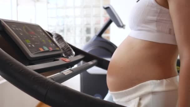 Здоровая беременность, люди и спортивная концепция. Беременная женщина ходит по беговой дорожке в фитнес-клубе . — стоковое видео
