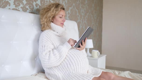 Zwangere vrouw met behulp van Tablet tijdens het rusten op bed in gezellige gebreide trui — Stockvideo