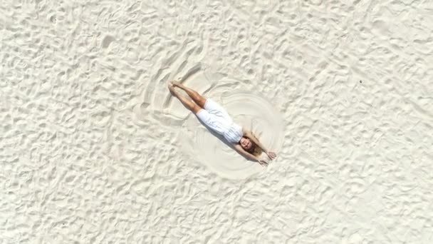 Γυναίκα με λευκό φόρεμα ξαπλωμένη στην παραλία κάνοντας τον άγγελο στην άμμο. Αεροσκάφος. — Αρχείο Βίντεο