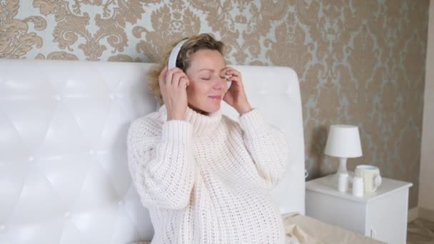 Молода вагітна жінка слухає музику і кладе навушники на живіт під час вагітності . — стокове відео