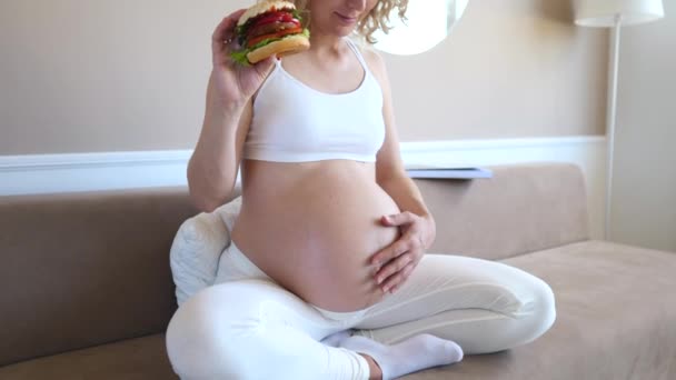 Πεινασμένη Έγκυος κορίτσι με χάμπουργκερ κάθεται στον καναπέ στο σπίτι. — Αρχείο Βίντεο