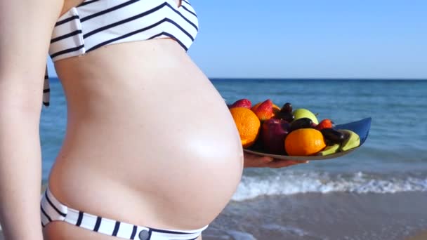 Κοντινό πλάνο της κοιλιάς της εγκύου γυναίκας στην παραλία δίπλα στη θάλασσα με φρέσκα φρούτα. — Αρχείο Βίντεο