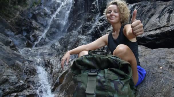 Женщина-туристка с рюкзаком показывает большие пальцы на водопаде — стоковое видео
