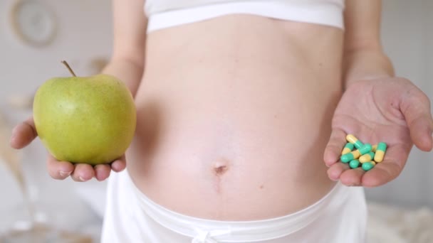 Die werdende Mutter hält Tabletten und grünen Apfel in der Hand. Nahaufnahme schwangerer Bauch. Pillen oder Früchte zur Wahl. — Stockvideo