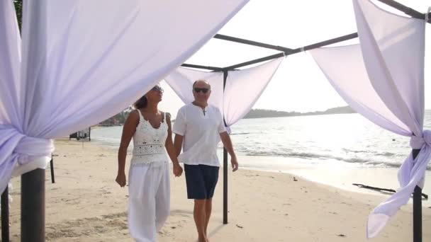Старшая пара в белой одежде, гуляющая по пляжу в летние каникулы — стоковое видео