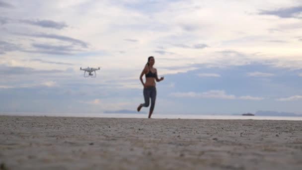 Vrouw rennend op het strand bij zonsondergang met vliegende drone — Stockvideo