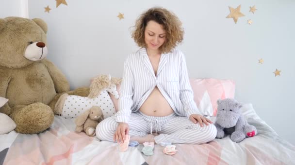 Joven mujer embarazada sonriente eligiendo calcetines para el futuro bebé usando pijamas en casa . — Vídeos de Stock