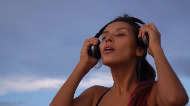 Jovem mulher multirracial em fones de ouvido ouvindo música na praia ao pôr do sol — Vídeo de Stock