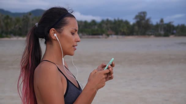 Garota de fitness com fones de ouvido usando smartphone depois de correr na praia — Vídeo de Stock