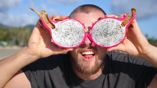 Grappige jongeman houdt roze pitaya fruit in plaats van ogen op het strand — Stockvideo