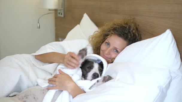Красивая женщина отдыхает в постели с собакой в халате — стоковое видео