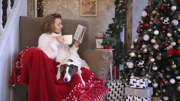 Νεαρή γυναίκα στο σπίτι διαβάζοντας βιβλίο με το σκυλί για τα Χριστούγεννα. Χειμερινές διακοπές έννοια. — Αρχείο Βίντεο