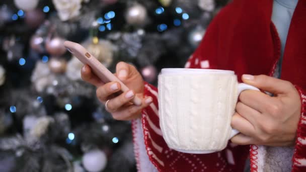 Primer plano de las manos femeninas con la taza usando el teléfono en el ambiente acogedor de la casa de Navidad — Vídeo de stock