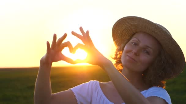 Дівчинка робить серцеподібну форму з руками на полі на заході сонця. — стокове відео