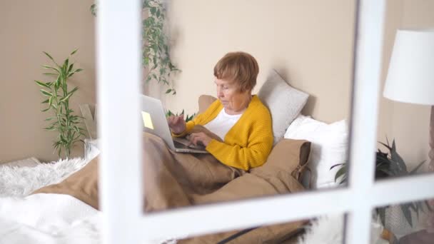 Ηλικιωμένη γυναίκα που χρησιμοποιεί φορητό υπολογιστή κάθεται στο κρεβάτι στο σπίτι. Τεχνολογία και οι ηλικιωμένοι έννοια. — Αρχείο Βίντεο