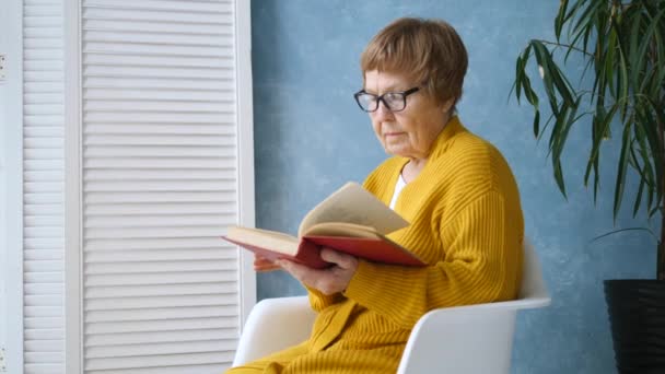 Ηλικιωμένη γυναίκα που διαβάζει ένα βιβλίο χαλαρώνοντας στο σπίτι. — Αρχείο Βίντεο