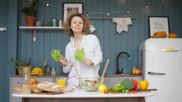 Fröhliche junge Frau tanzt mit grünem Salat in Küche — Stockvideo