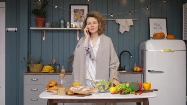 Mujer joven hablando por teléfono celular en la cocina mientras cocina comida saludable . — Vídeos de Stock