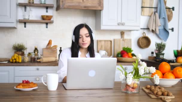Junge brünette Frau mit Laptop trägt Kopfhörer und frühstückt in der Küche. — Stockvideo