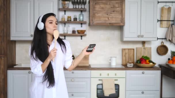 Mulher jovem com telefone celular cantando na cozinha usando fones de ouvido — Vídeo de Stock