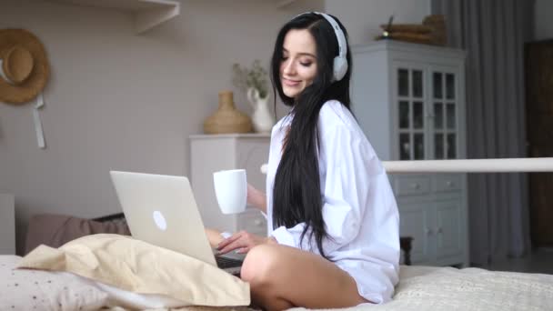 ノートパソコンで働く女の子とヘッドフォンをして朝コーヒーを飲む. — ストック動画