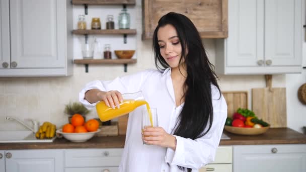 Mulher morena bonita derramando suco de laranja em vidro na cozinha pela manhã — Vídeo de Stock