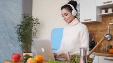 Mutfakta dizüstü bilgisayar kullanan, kulaklık takan, gülümseyen genç bir kadın.