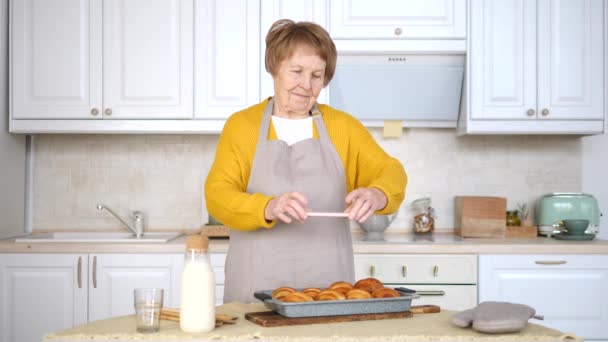 Mulher sênior tirando foto de pastelaria recém-assada usando celular na cozinha . — Vídeo de Stock