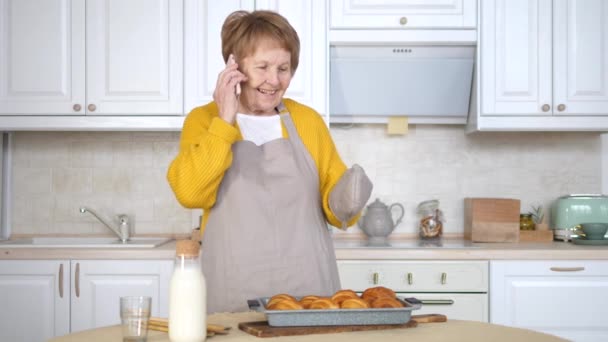 Oudere vrouw spreken op de mobiele telefoon tijdens het bakken in de keuken — Stockvideo