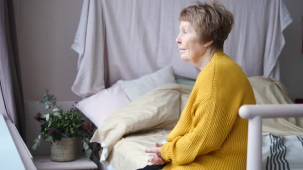 Femme âgée réfléchie assise sur son lit regardant par la fenêtre — Video