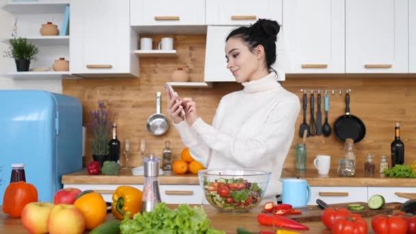 Jeune femme utilisant le téléphone portable dans la cuisine tout en cuisinant . — Video