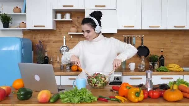 Kulaklık takan, salata yapan ve mutfakta dizüstü bilgisayara bakan güzel bir kadın. — Stok video