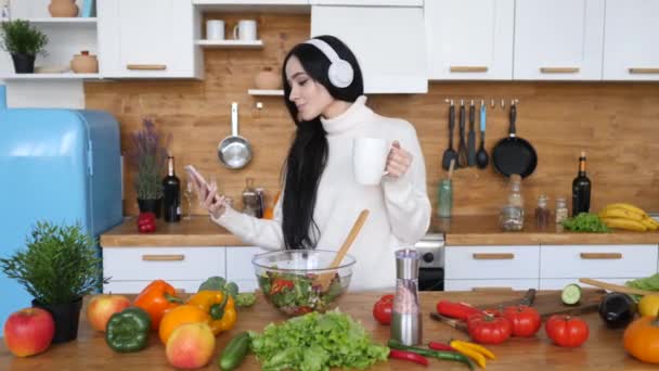 スマートフォンを使い、ヘッドフォンを装着したキッチンで踊る女性. — ストック動画